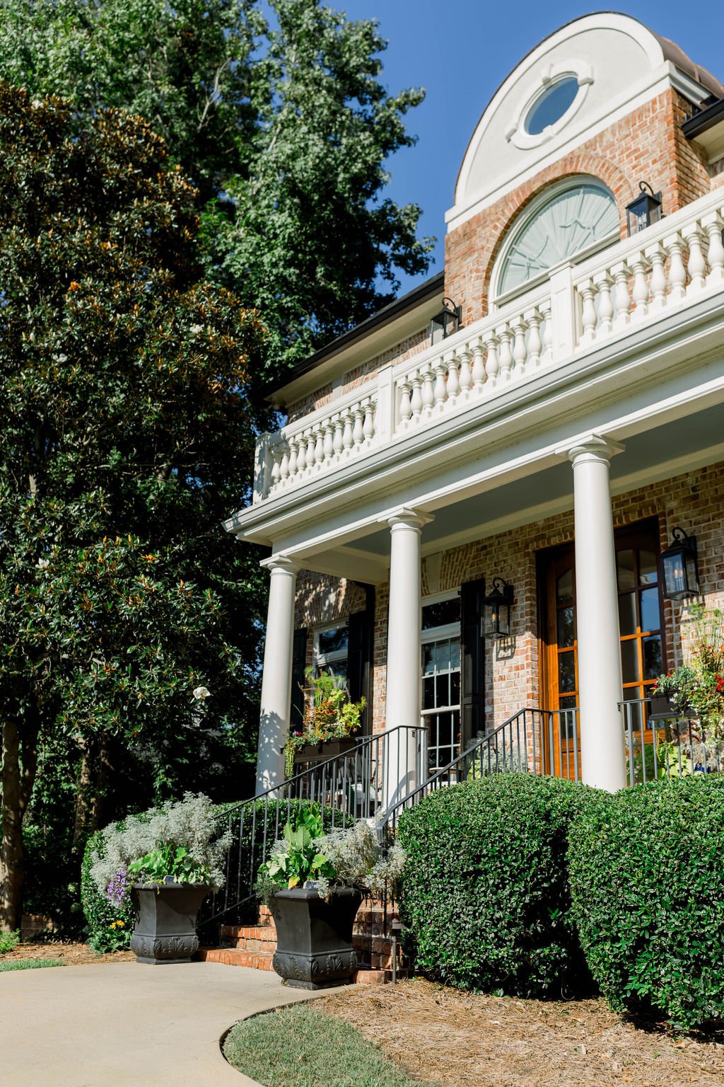 Front porch of a brick house with white columns, a balcony with a white railing, and large windows. There are potted plants on the stairs and greenery around the house, including bushes and tall trees, under a clear blue sky.