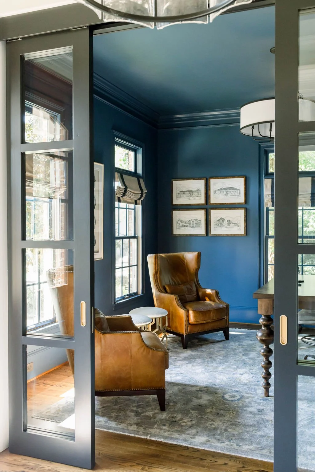 Interior view of a cozy room with blue walls, two large windows, a leather armchair, and framed architectural sketches on the wall, as seen through a glass-paneled sliding door.