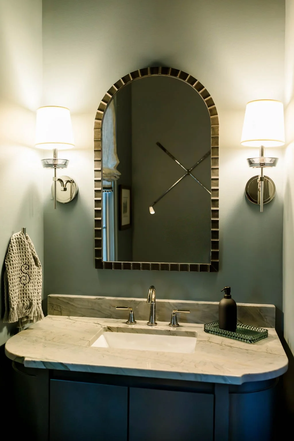 Bathroom with an oval mirror, two wall sconces with lamps, a marble sink with a brushed nickel faucet, a black soap dispenser, and a monogrammed towel hanging on the wall.