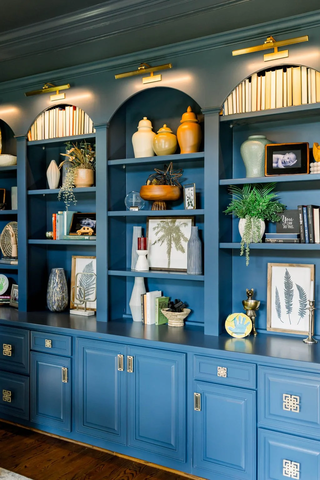 A blue built-in bookshelf with decorative vases, framed art, potted plants, books, and various decorative objects, illuminated by gold picture lights.