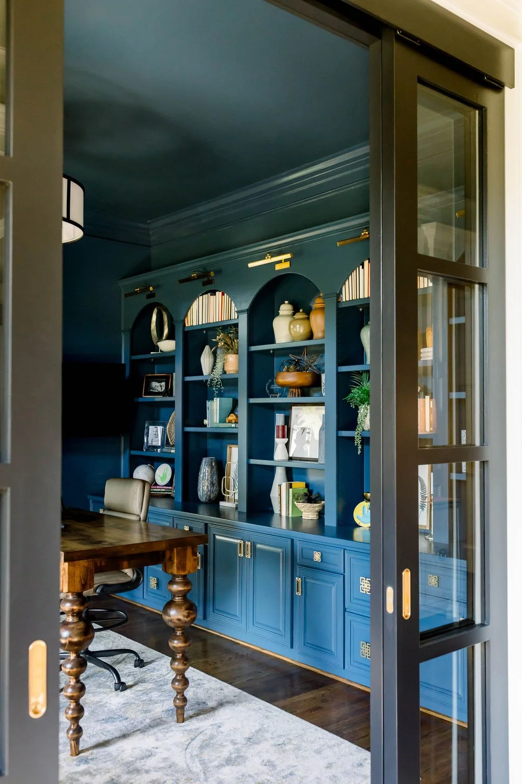 A room with blue built-in shelves filled with decorative vases, plants, and framed photos, visible through a sliding glass door from an adjacent room with a wooden dining table and beige chair.