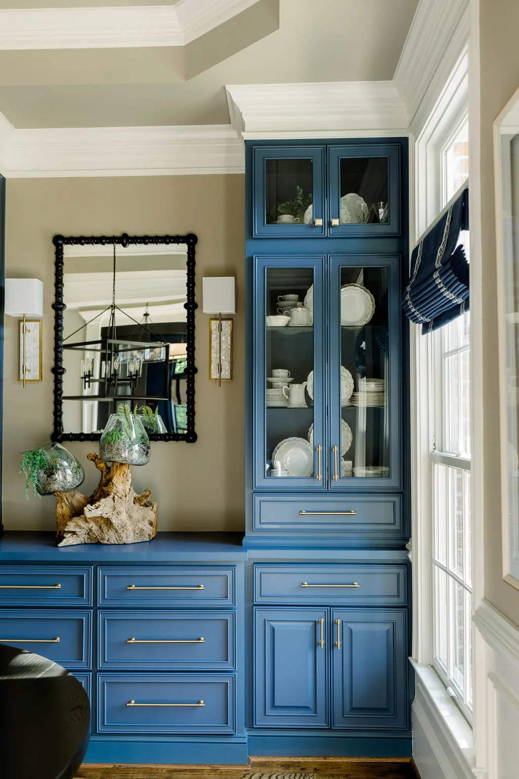 Blue built-in china cabinet with glass doors displaying white dishware next to a large window with blue Roman shades in a dining room.