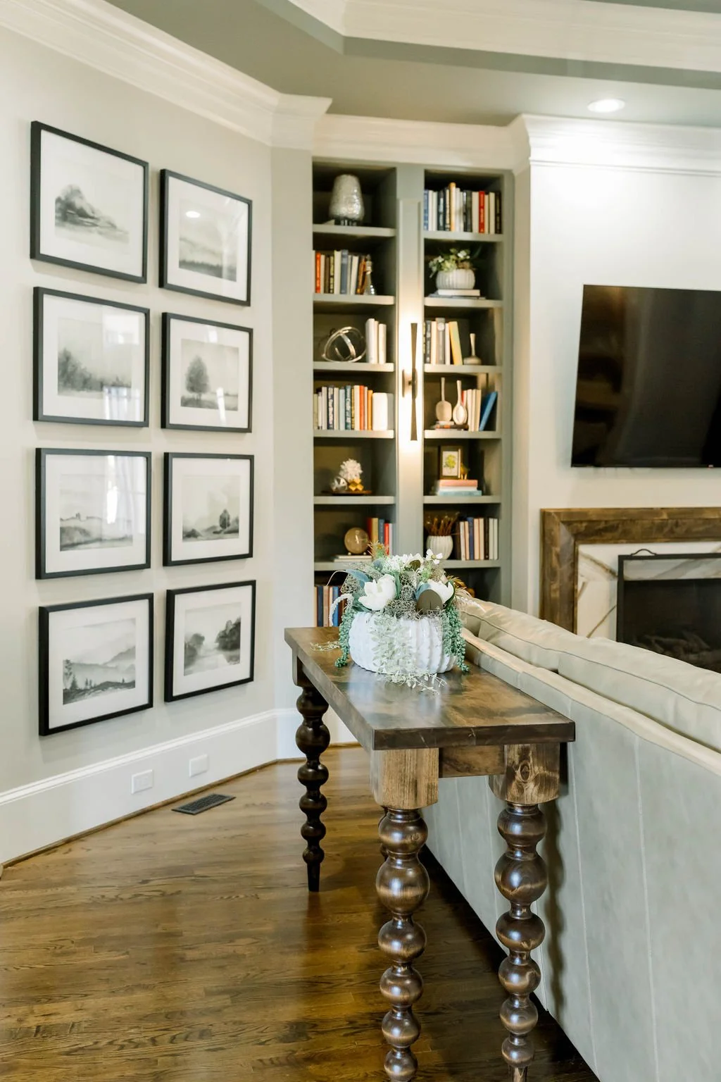 Living room with a framed black-and-white landscape art on the wall, a wooden console table with a floral arrangement, a built-in bookshelf filled with books, a fireplace with a wooden mantel, and a mounted flat-screen TV.