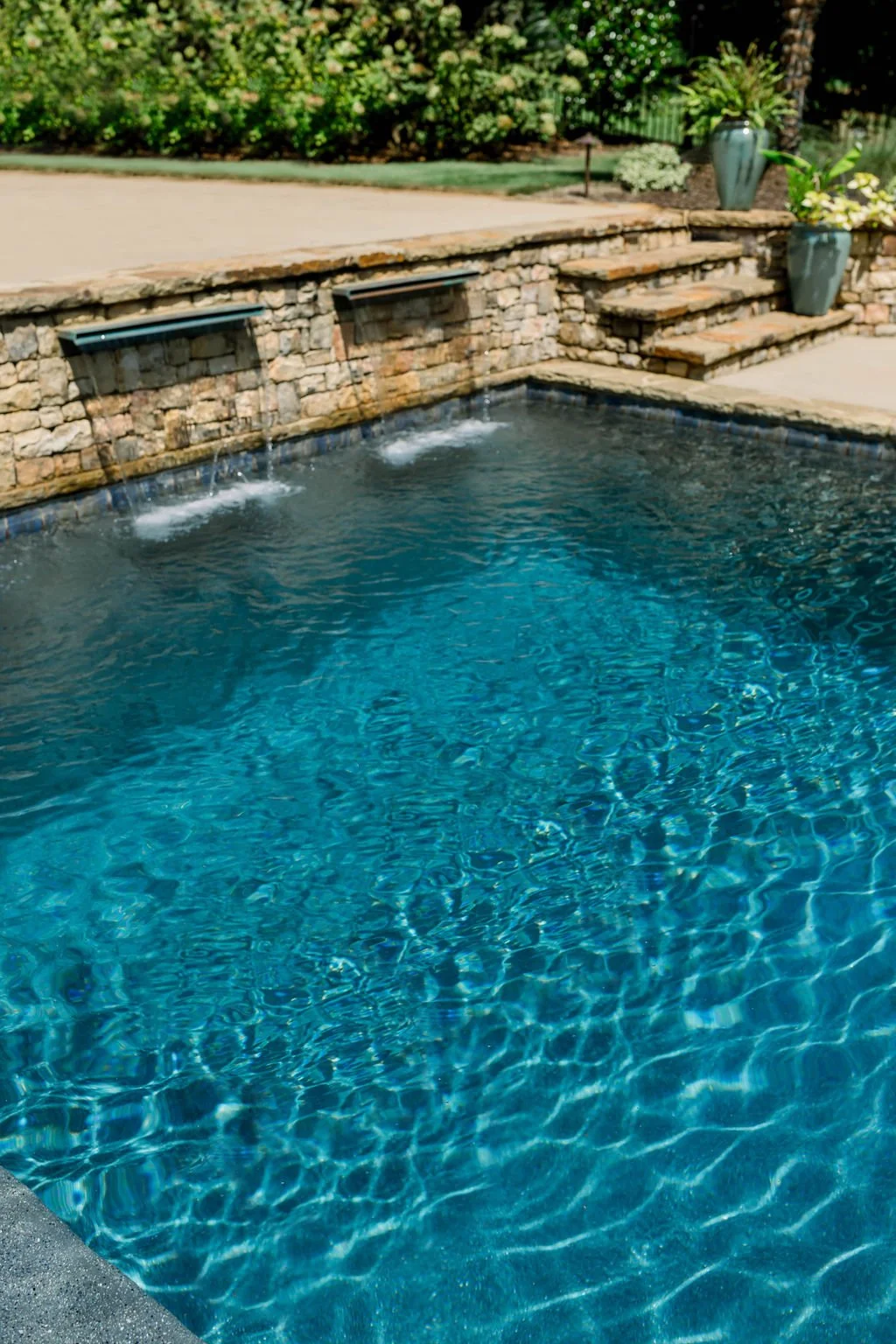 Swimming pool with water features on a stone wall, surrounded by green bushes and potted plants.