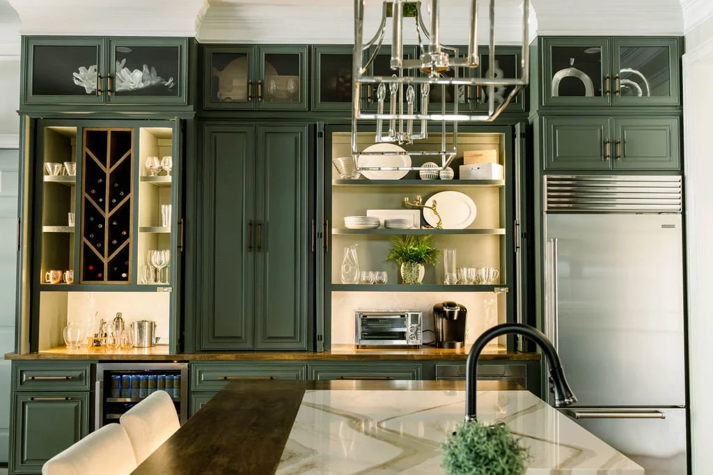 Modern kitchen with dark green cabinets, open shelves with glassware, wine storage, and built-in refrigerator, island with marble countertop, black faucet, and chandelier.