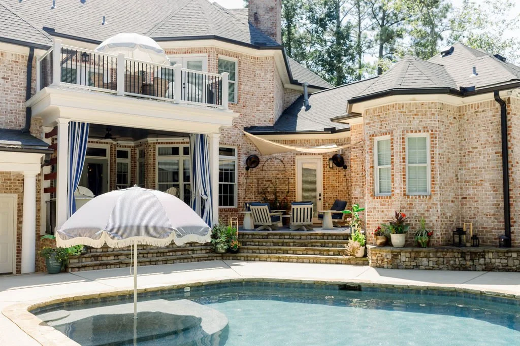 Backyard with a swimming pool, patio furniture, an umbrella, potted plants, and a multi-story brick house with a balcony and large windows.