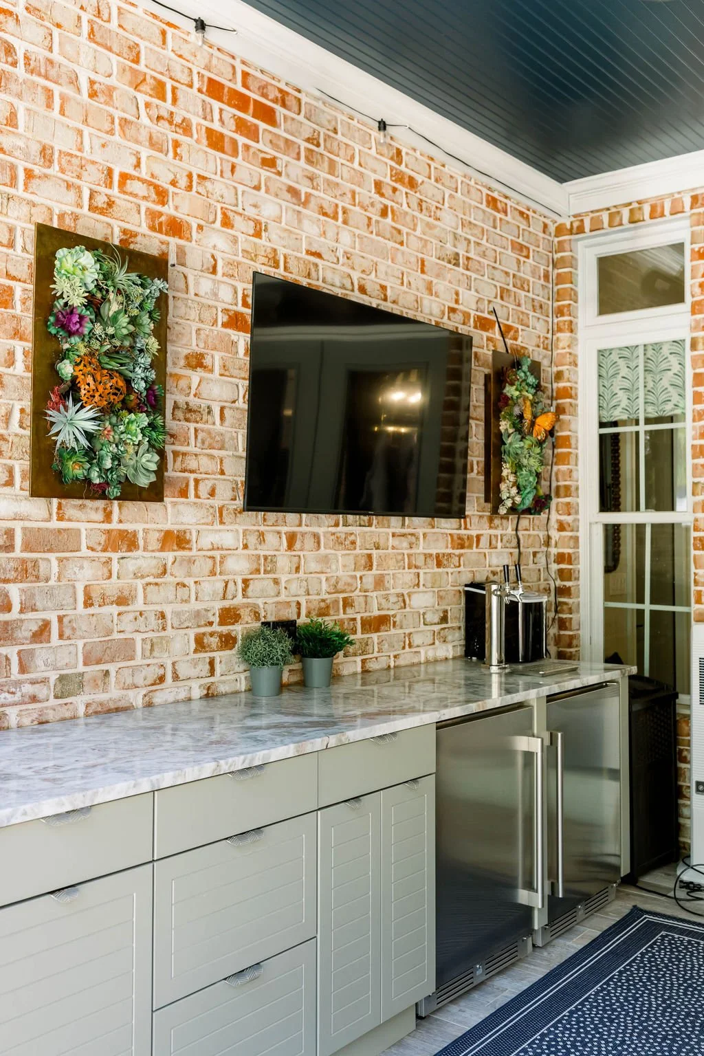 Indoor outdoor kitchen area with a brick wall, a mounted flat-screen TV, two vertical plant displays, a marble countertop, small potted plants, a mini refrigerator, and a window with decorative blinds.