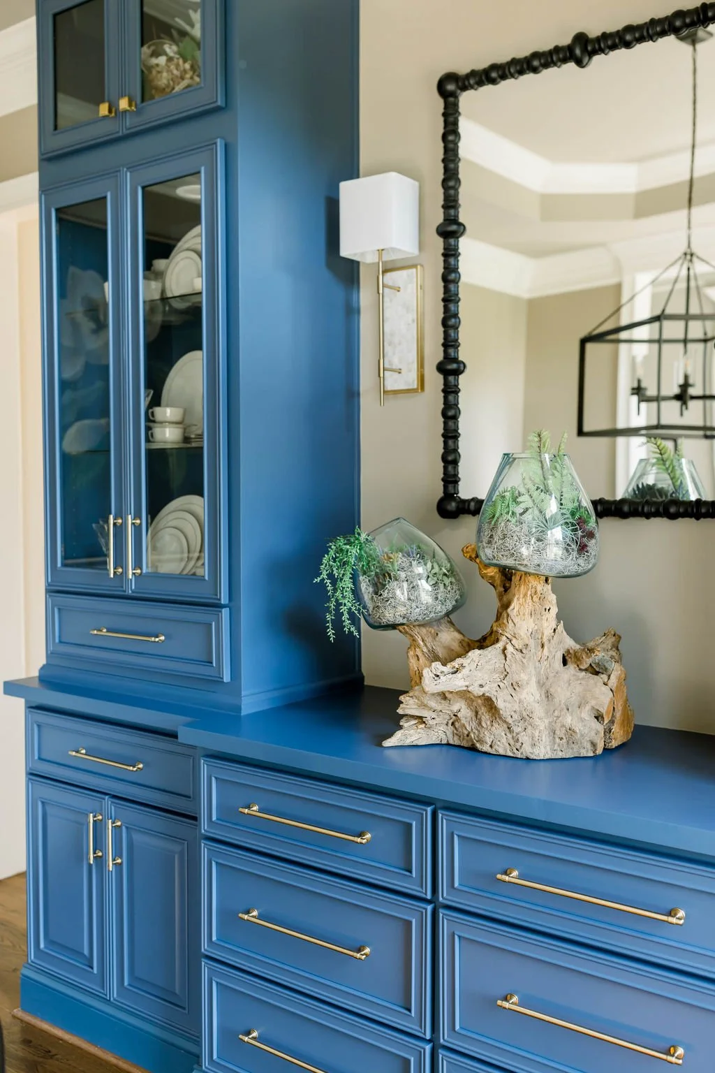 A blue china cabinet with glass doors displaying white dishes and bowls, next to a wire frame mirror and a decorative wooden centerpiece with glass terrariums containing plants.