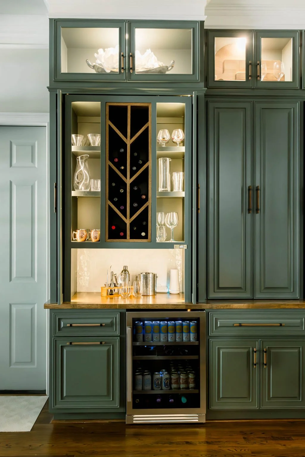 Built-in cabinetry with glass-front upper cabinets and a wine cooler, featuring glassware, a wooden countertop, and a mini fridge at the bottom, in a kitchen.