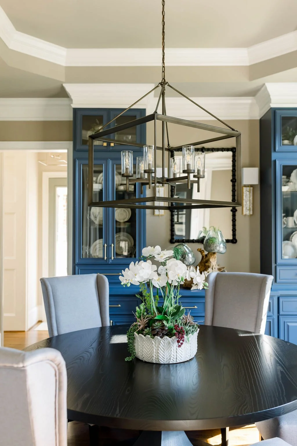 A dining room with a round black wooden table, white floral centerpiece, beige upholstered chairs, blue built-in cabinets with glass doors, and a black metal chandelier hanging from the ceiling.
