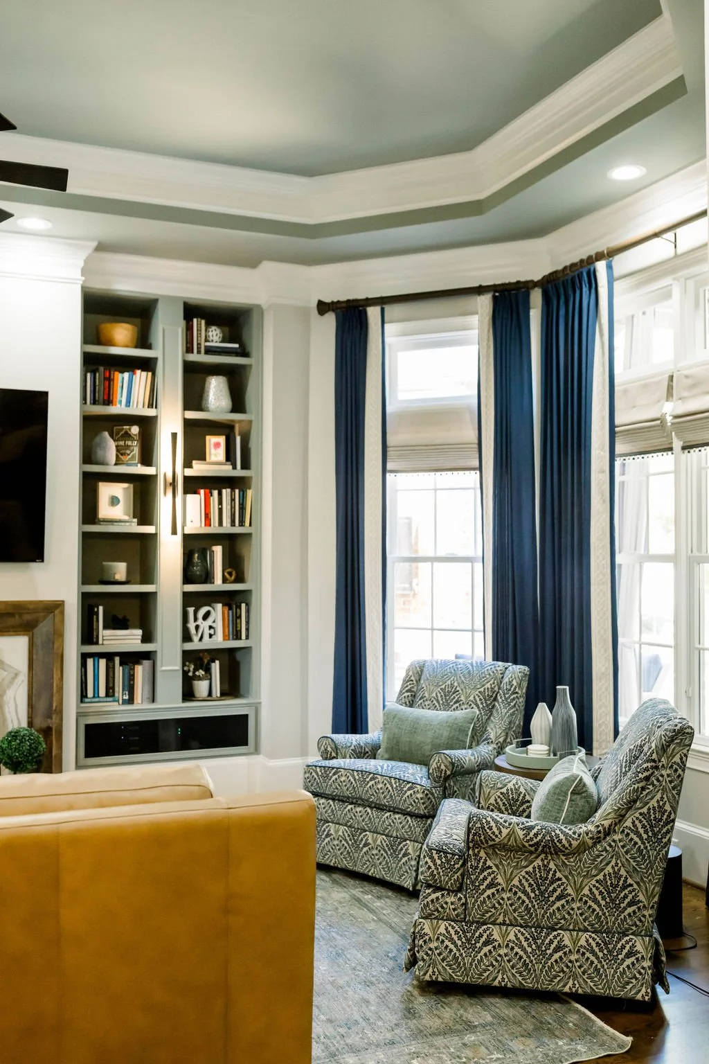 Living room with gray walls, built-in bookshelf, blue curtains, patterned armchairs, and a tan sofa.