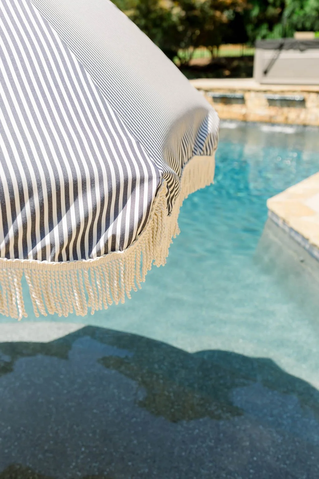 Close-up of a blue and white striped patio umbrella with beige fringe near a swimming pool in sunny outdoor setting.