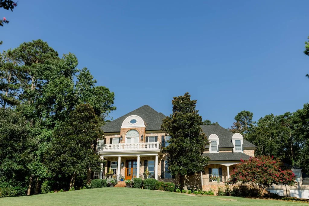 Large brick house with a gray cone-shaped roof, surrounded by green trees and a grassy lawn, under a clear blue sky.