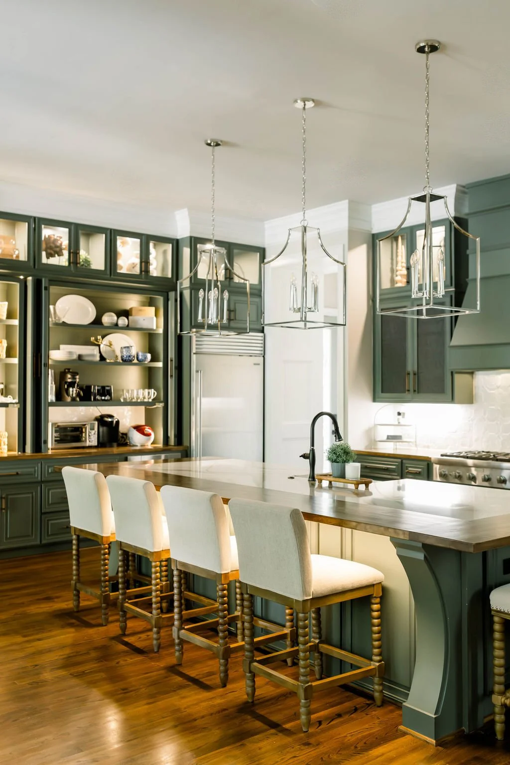 Bright kitchen with dark green cabinets, white countertops, pendant lighting, and a wooden island with white barstools.