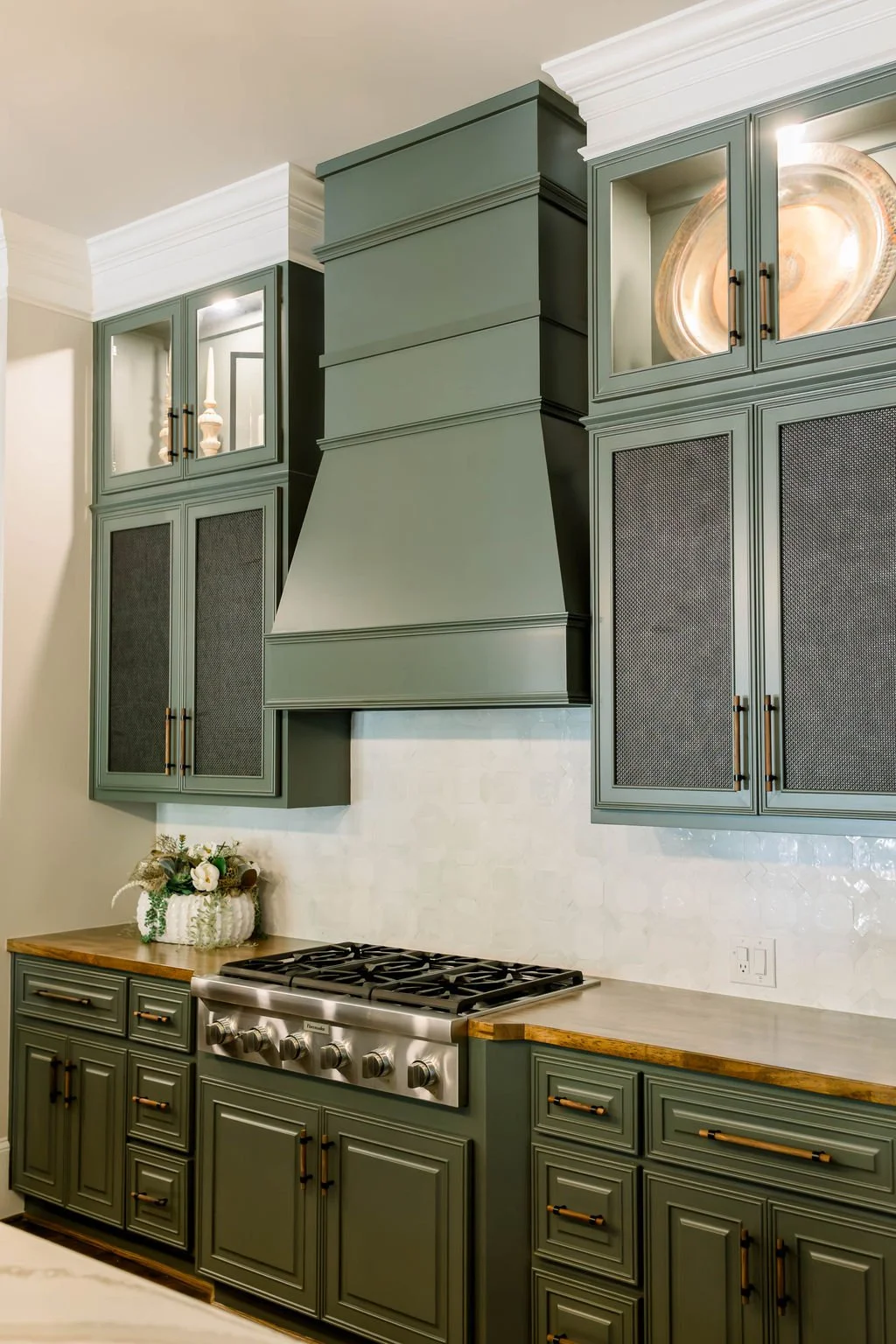 Green kitchen cabinets with black handles, a stainless steel stove, and decorative items like a floral arrangement and a large dish on a shelf.