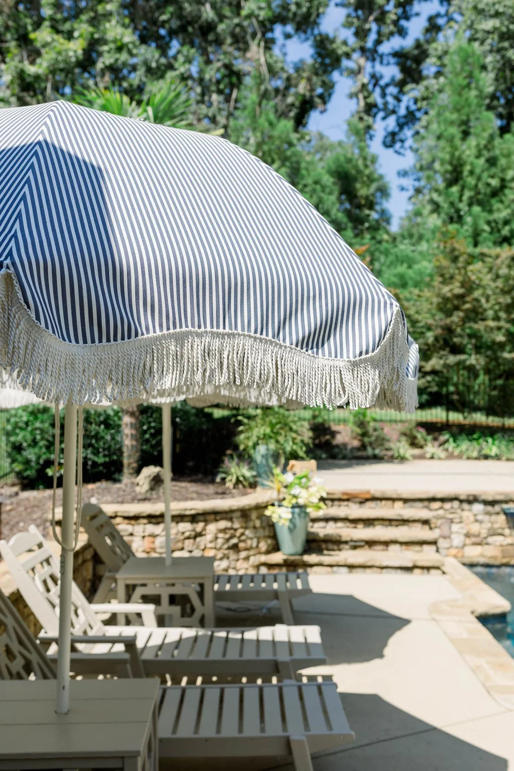 Poolside scene with striped umbrella, lounge chairs, and surrounding greenery.