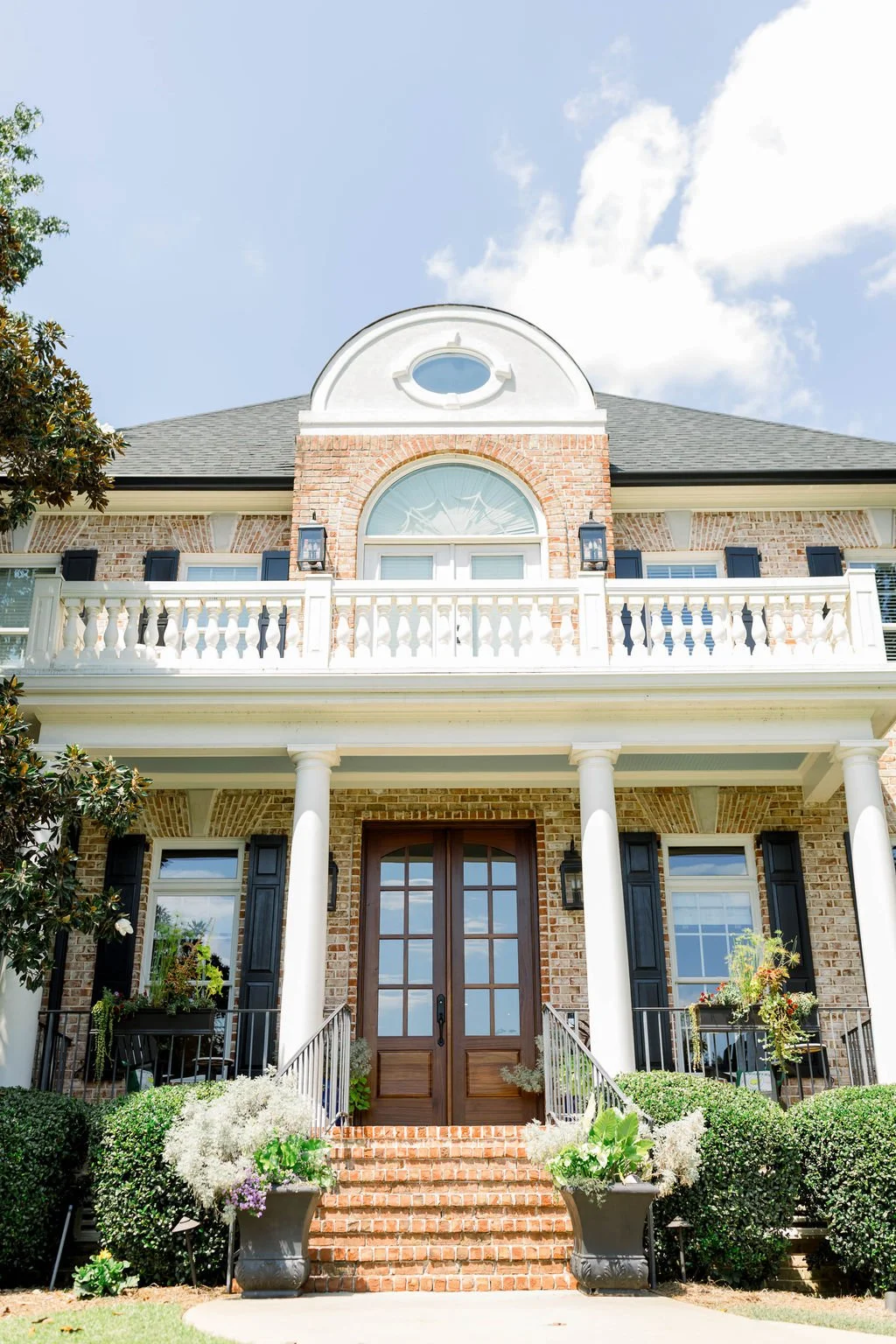 Front view of a large brick house with white columns and black shutters, featuring a balcony and front door with potted plants and bushes.