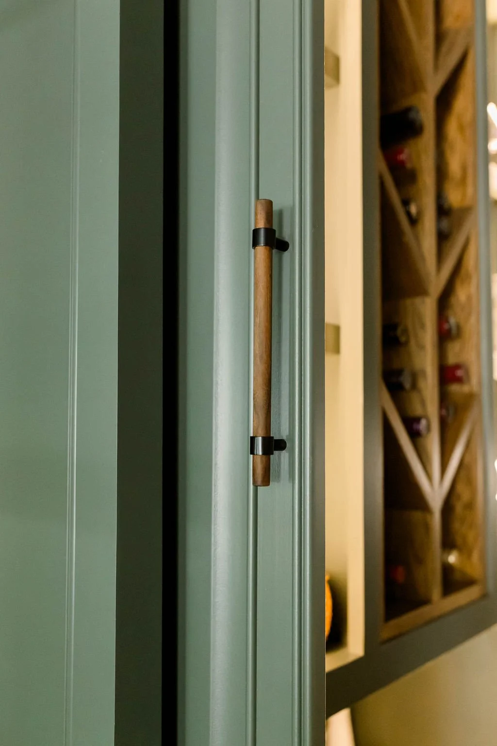 Close-up of a green cabinet door with a wooden and black handle, with a wine rack filled with bottles visible in the background.