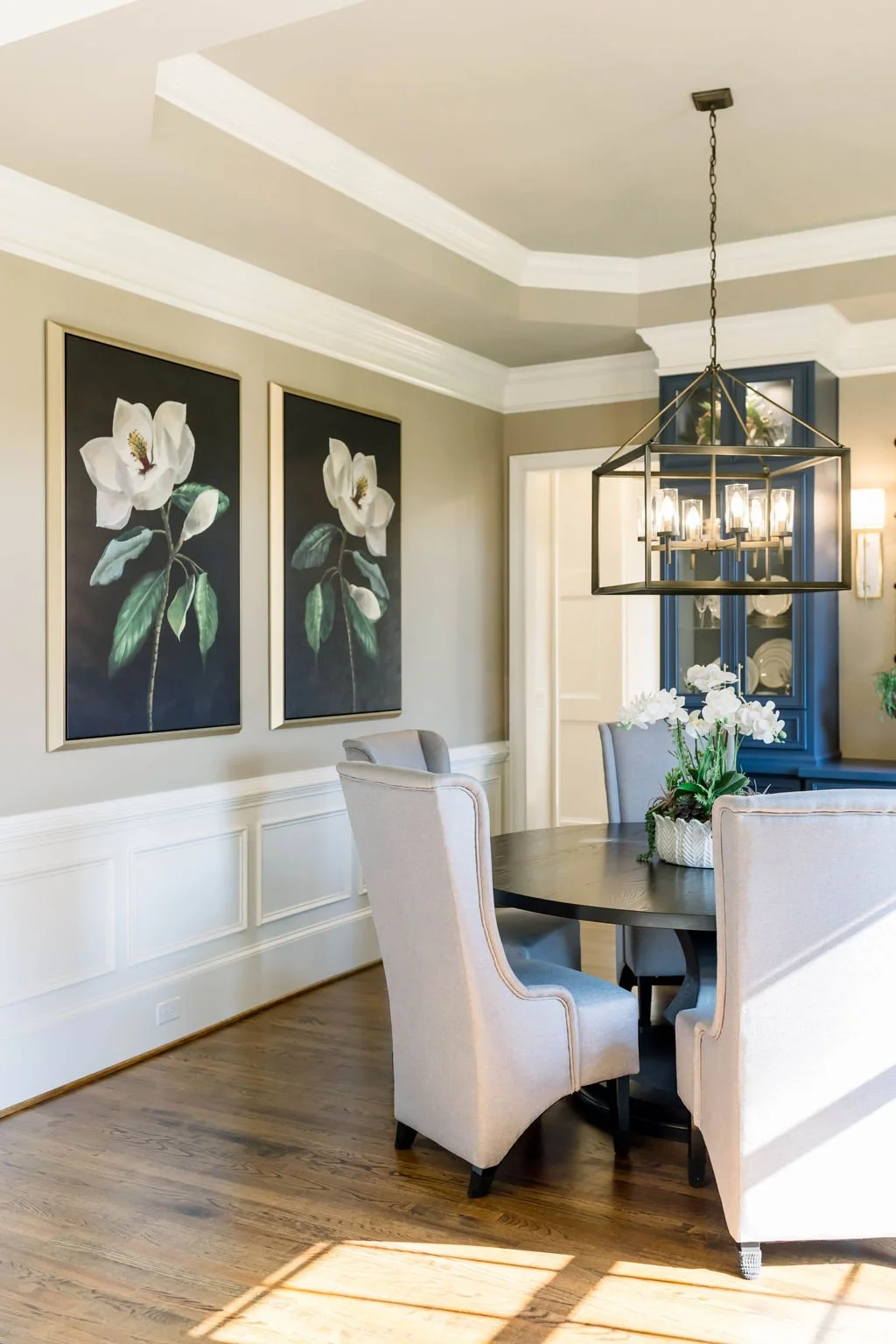 Modern dining room with beige walls, white crown molding, and hardwood floors. Features a round dark wood table with four upholstered beige chairs. A black chandelier hangs above the table, and a blue china cabinet is in the background. There are two