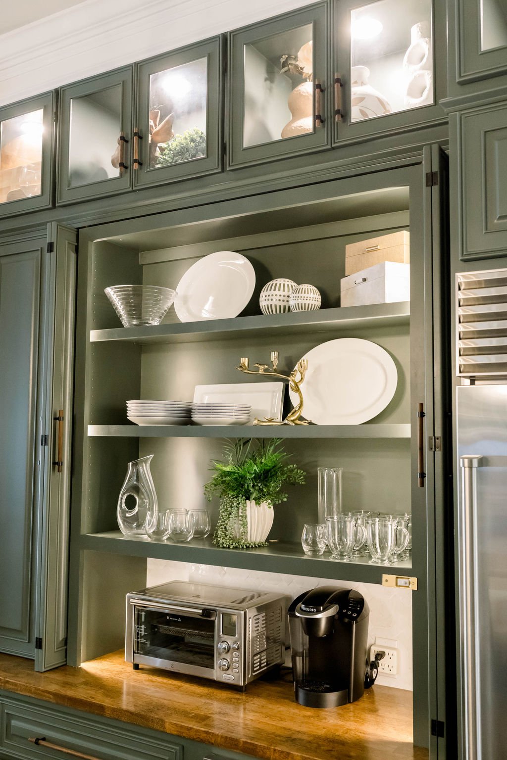 Open green kitchen cabinet displaying dishes, glassware, and decorative items, with a wooden countertop holding a toaster oven and coffee machine.