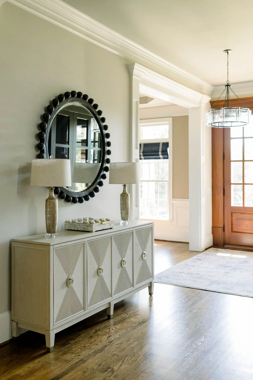 Entryway with a white sideboard, two lamps, a mirror, and a chandelier in a well-lit room with hardwood floors and a front door.