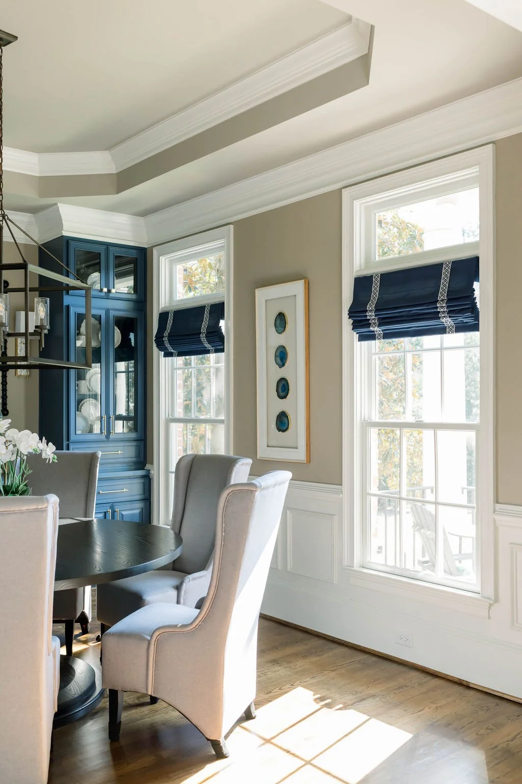 A bright dining room with two large windows with white frames and blue Roman shades, beige walls with white wainscoting, a black round dining table with beige upholstered chairs, a blue built-in china cabinet with glass doors, and a framed art piece 