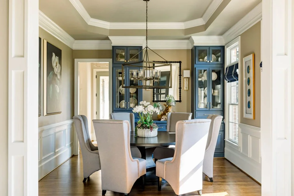 A dining room with a round table surrounded by six upholstered chairs. There is a floral centerpiece on the table, blue cabinets with glass doors displaying plates and glassware, a large rectangular chandelier hanging above the table, and framed artw