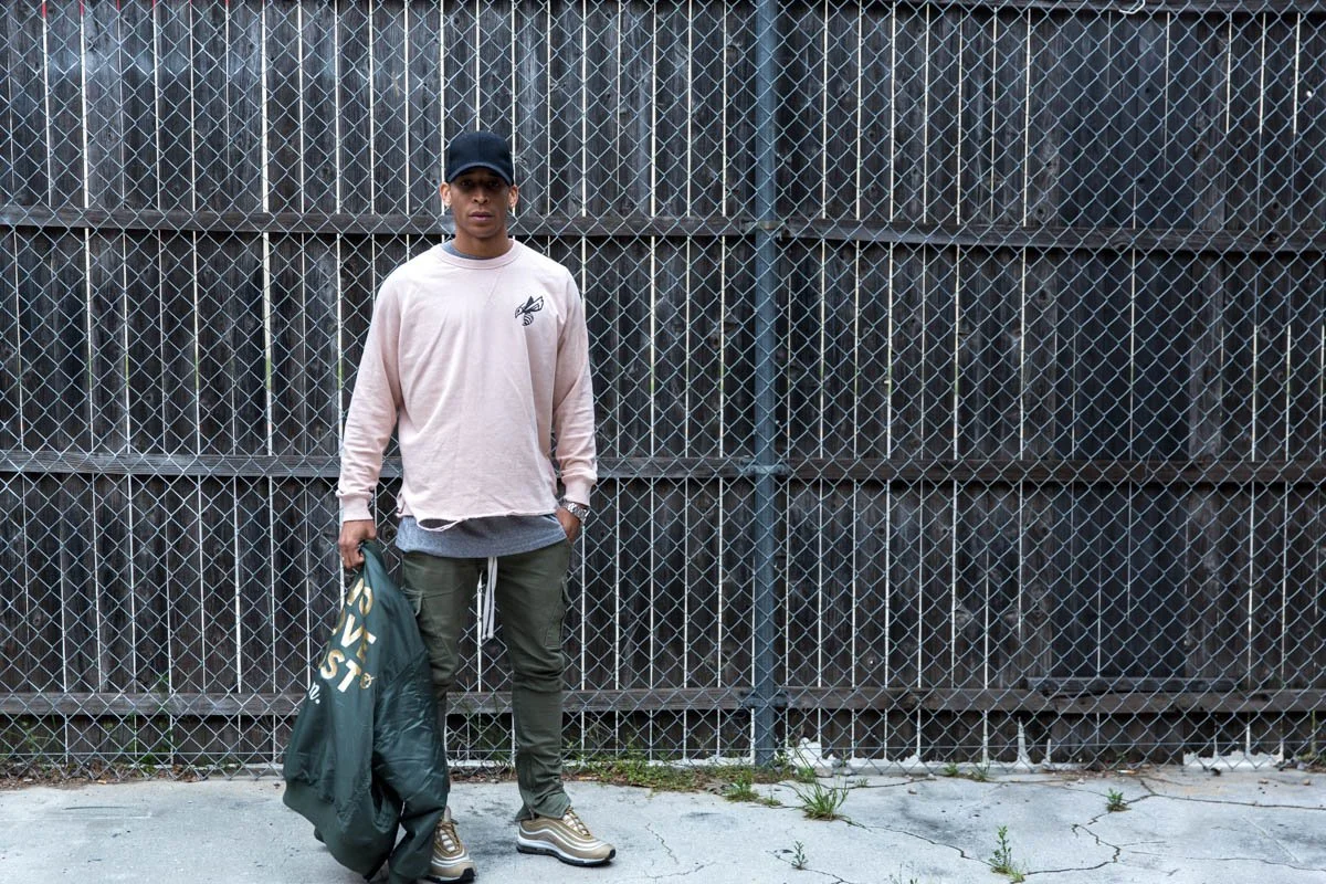 A young man stands in front of a chain-link fence, wearing a beige long-sleeve shirt, dark green pants, and sneakers, holding a bag in his left hand.