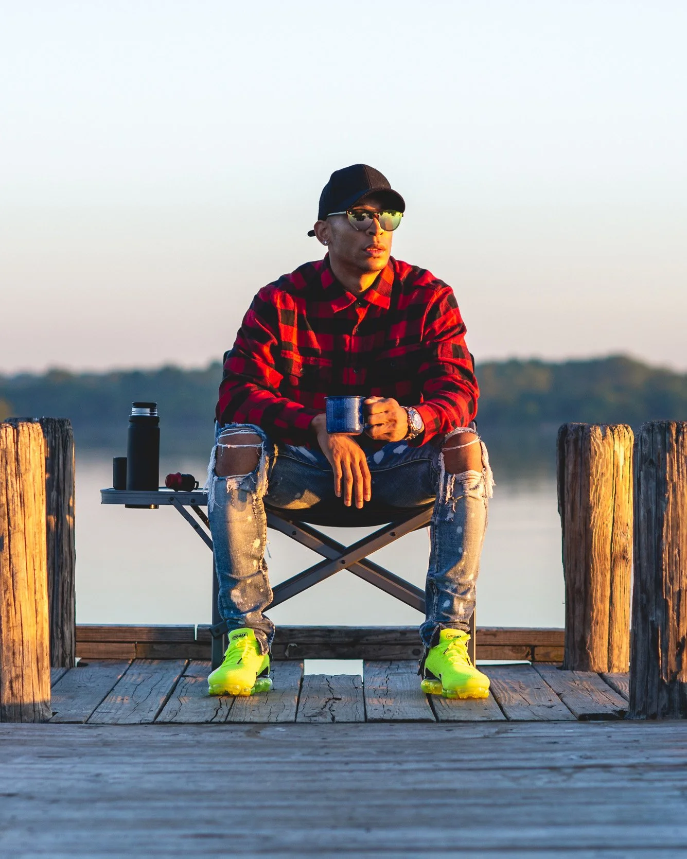 A man sitting on a stool on a wooden dock by the water, wearing a black cap, reflective sunglasses, a red and black checkered shirt, ripped jeans, and bright yellow neon green, holding a mug.