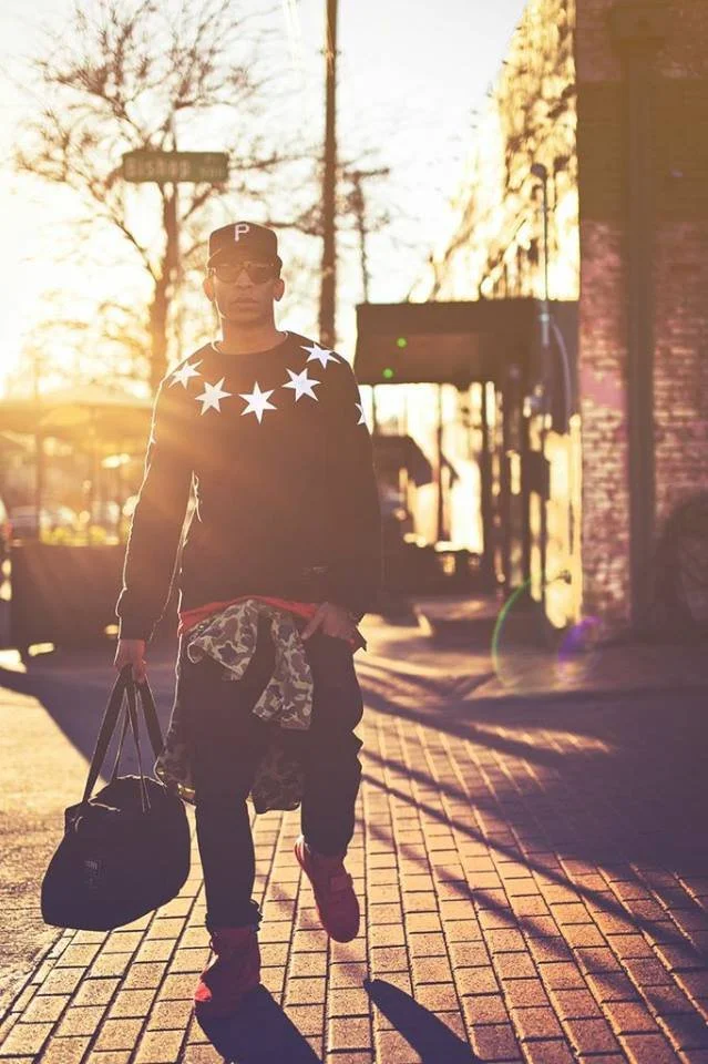 A young man walking on a city sidewalk during sunset, carrying a bag, wearing sunglasses, a cap with a 'P', a star-patterned sweater, and camouflage jacket tied around his waist.