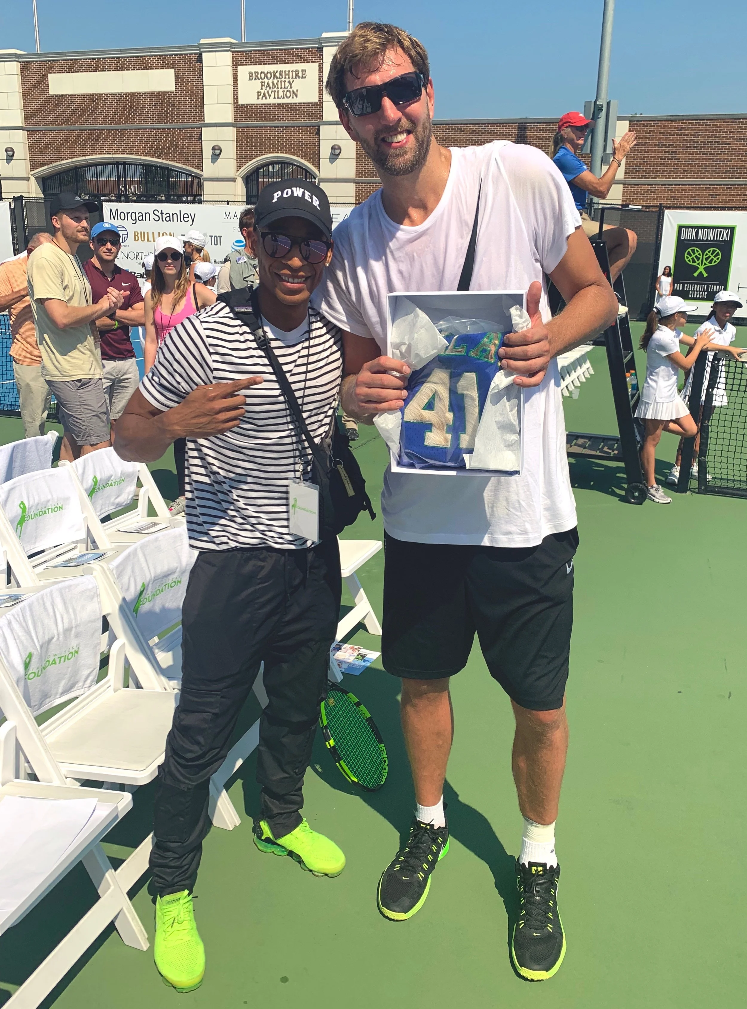 Leon McKenzie standing next to Dirk Nowitzki. They are together at SMU Tennis Complex, smiling, with Dirk holding a MIEL Y NOIR gift box which included a customized jersey and other items.