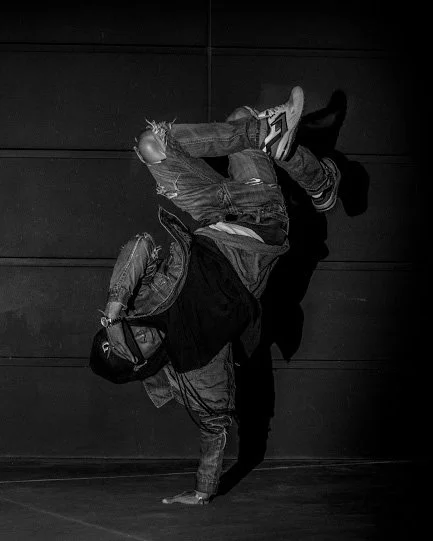 Person performing a breakdance move on the floor while holding a football, with a shadow cast on a wall in a dark setting.