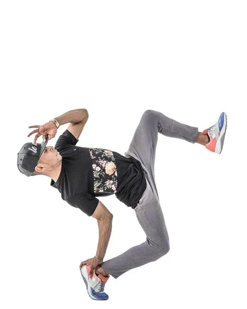 Person in a rotational pose adjusting sunglasses, wearing a black shirt with floral design, gray sweatpants, and colorful sneakers.