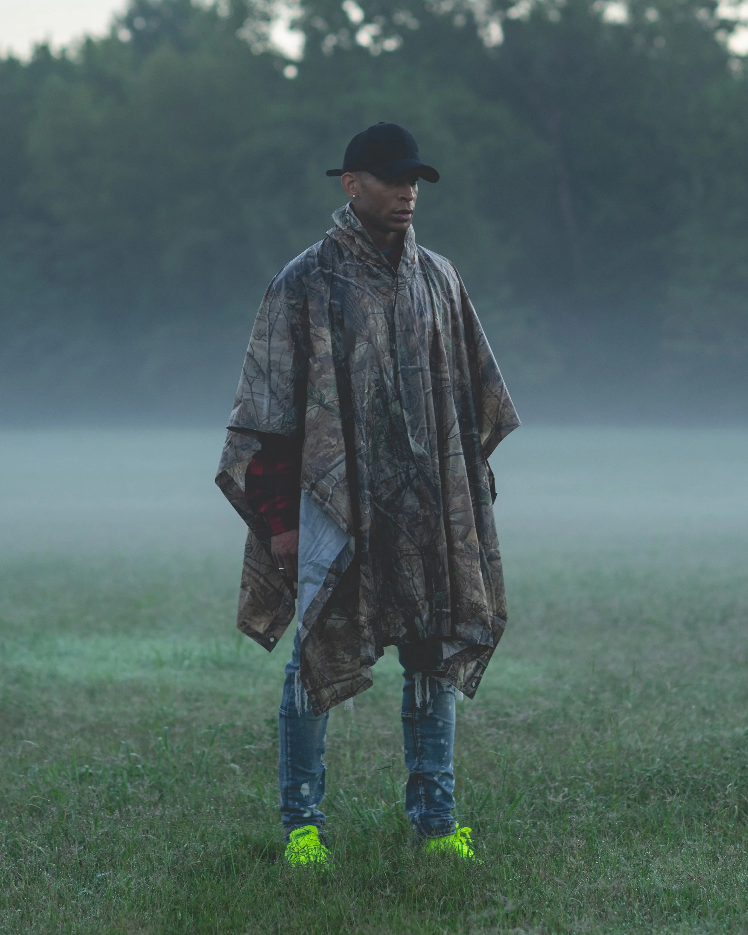 A person standing in a foggy outdoor setting wearing a camouflage poncho, a black cap, ripped jeans, and neon yellow shoes.