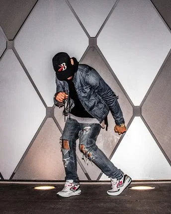 A person wearing a black cap, black face mask, denim jacket, ripped jeans, and white Sneakers, standing in an elevator with geometric wall panels.