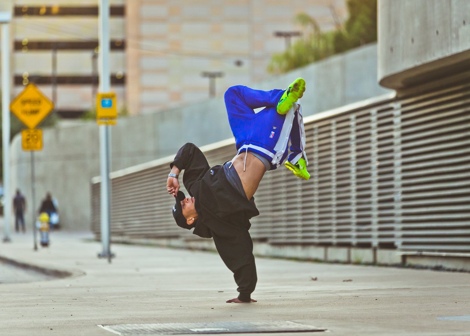 A man performing a handstand street dance move outdoors on a city sidewalk, wearing black hoodie, blue jacket with white stripes, black cap, and bright yellow shoes.
