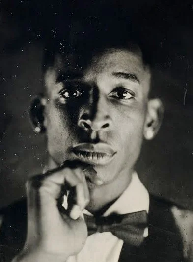 Black and white pinhole camera portrait of a young man in formal attire, with a bow tie, holding his chin with one hand, looking directly at the camera.