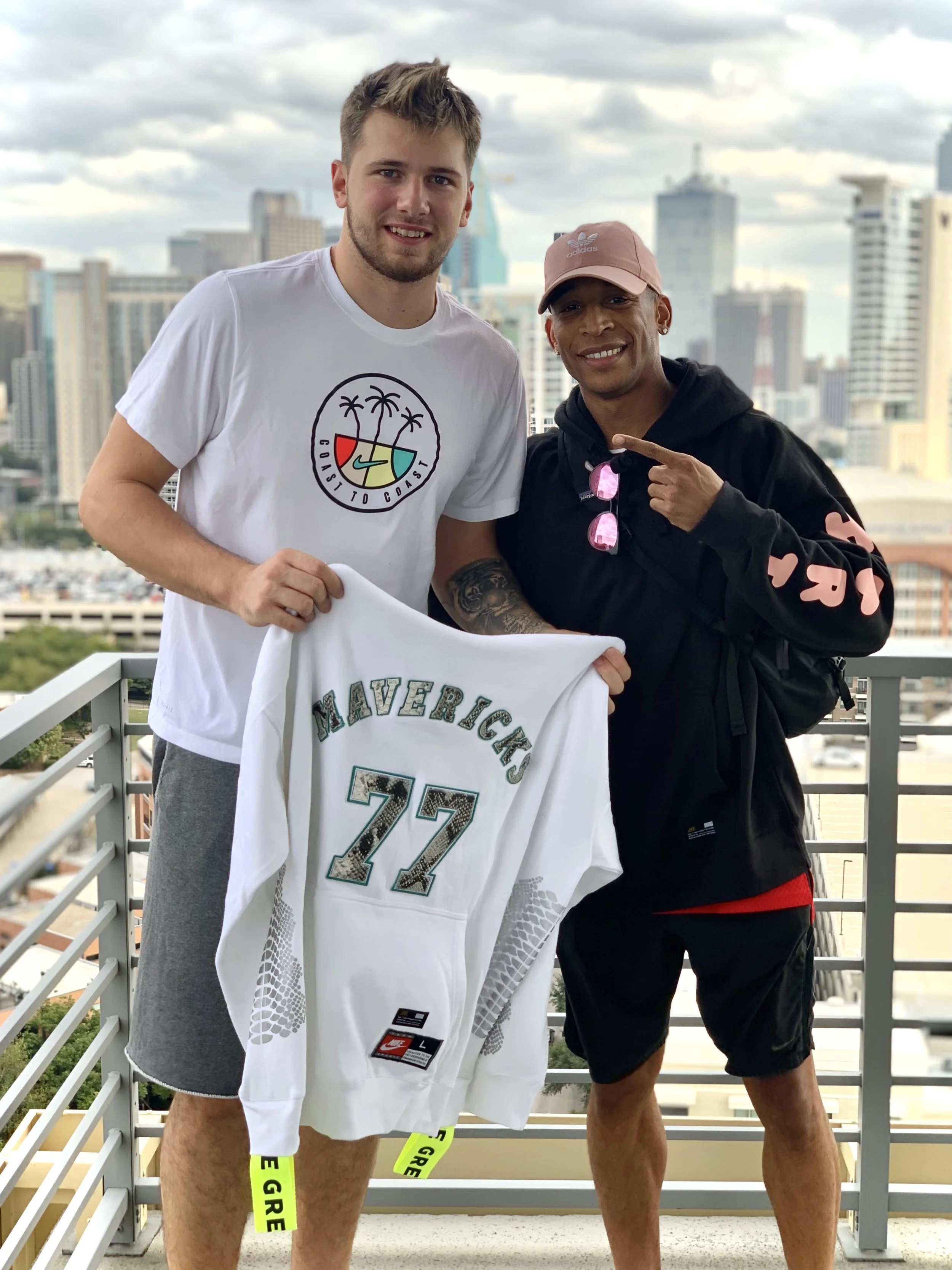 Leon McKenzie is standing next to Luka Doncic, standing on a balcony with a cityscape in the background after gifting him a MIEL Y NOIR package. They are smiling and holding a customized Dallas Mavericks hoodie.