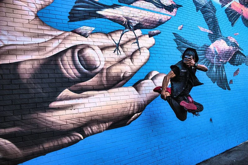 A person is jumping in front of a mural on a brick wall that depicts a large hand holding a small bird, with other birds flying in the background.