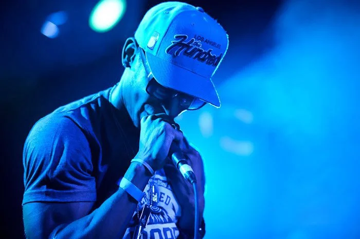 A man wearing a gray baseball cap with 'Los Angeles' and 'Ford' embroidered on it, a gray T-shirt, and sunglasses, holding a microphone on stage with blue lighting.