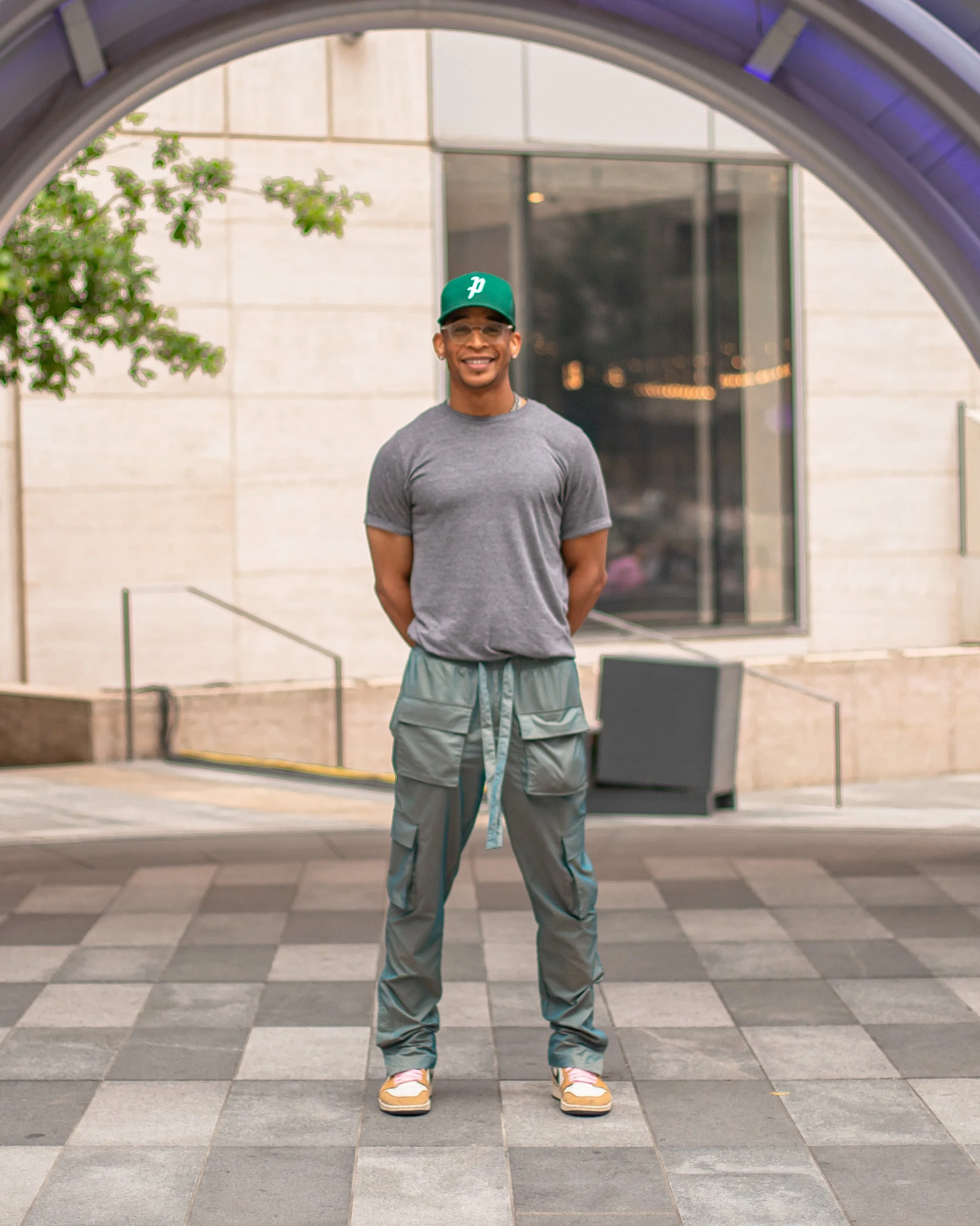Leon McKenzie, 'PRO' stands outdoors at the AT&T Discovery Zone in downtown Dallas, smiling, wearing a gray t-shirt, iridescent green drawstring pants, Rookie Of The Year Jordan 1's, and a green baseball cap.