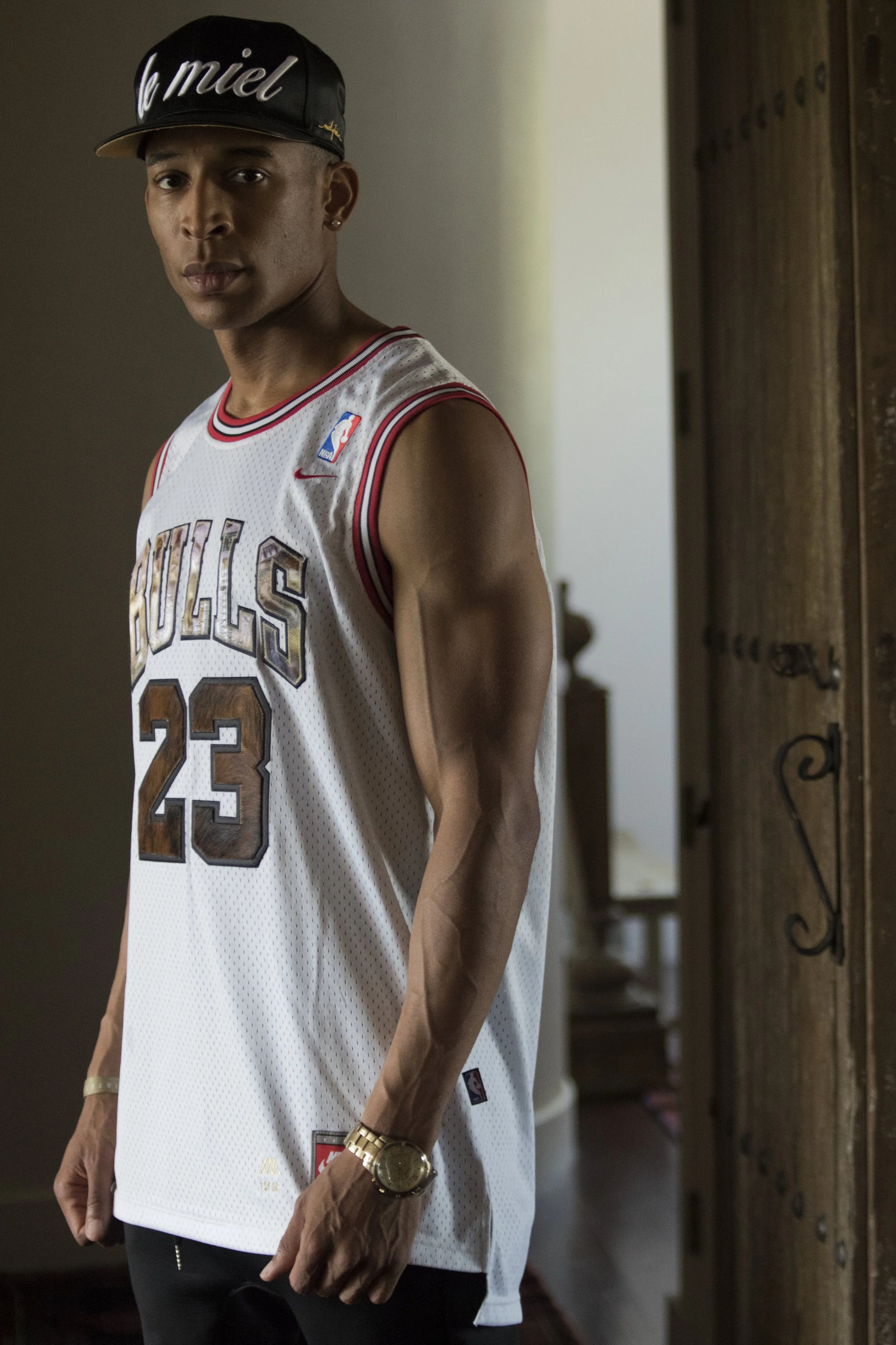 A young man wearing a white Chicago Bulls basketball jersey with the number 23, a black cap with 'le miel' written on it, a gold watch, a bracelet, and earrings, standing indoors near a wooden door, looking at the camera.