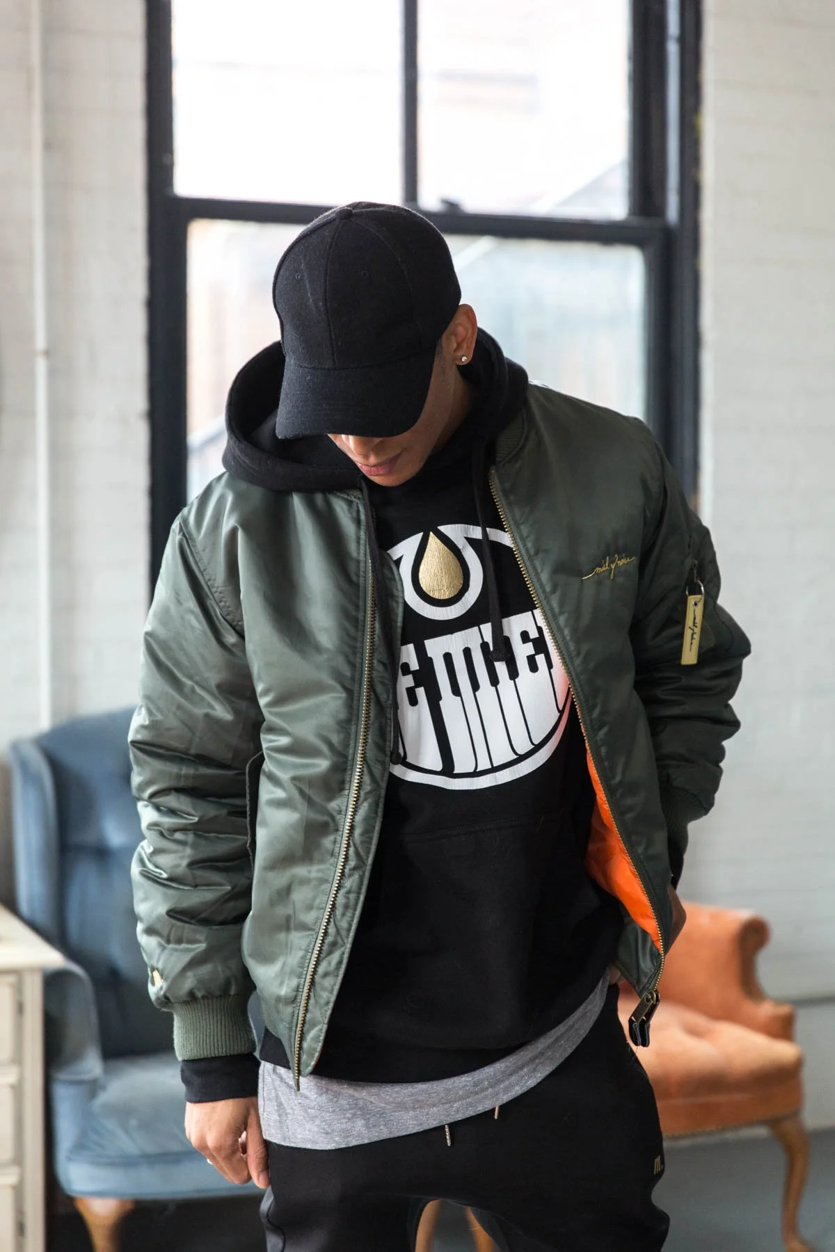 A man wearing a black baseball cap, black hoodie, olive green bomber jacket with a gold zipper, and dark pants, standing indoors near large windows with brick and white walls, and a tan vintage-style armchair in the background.