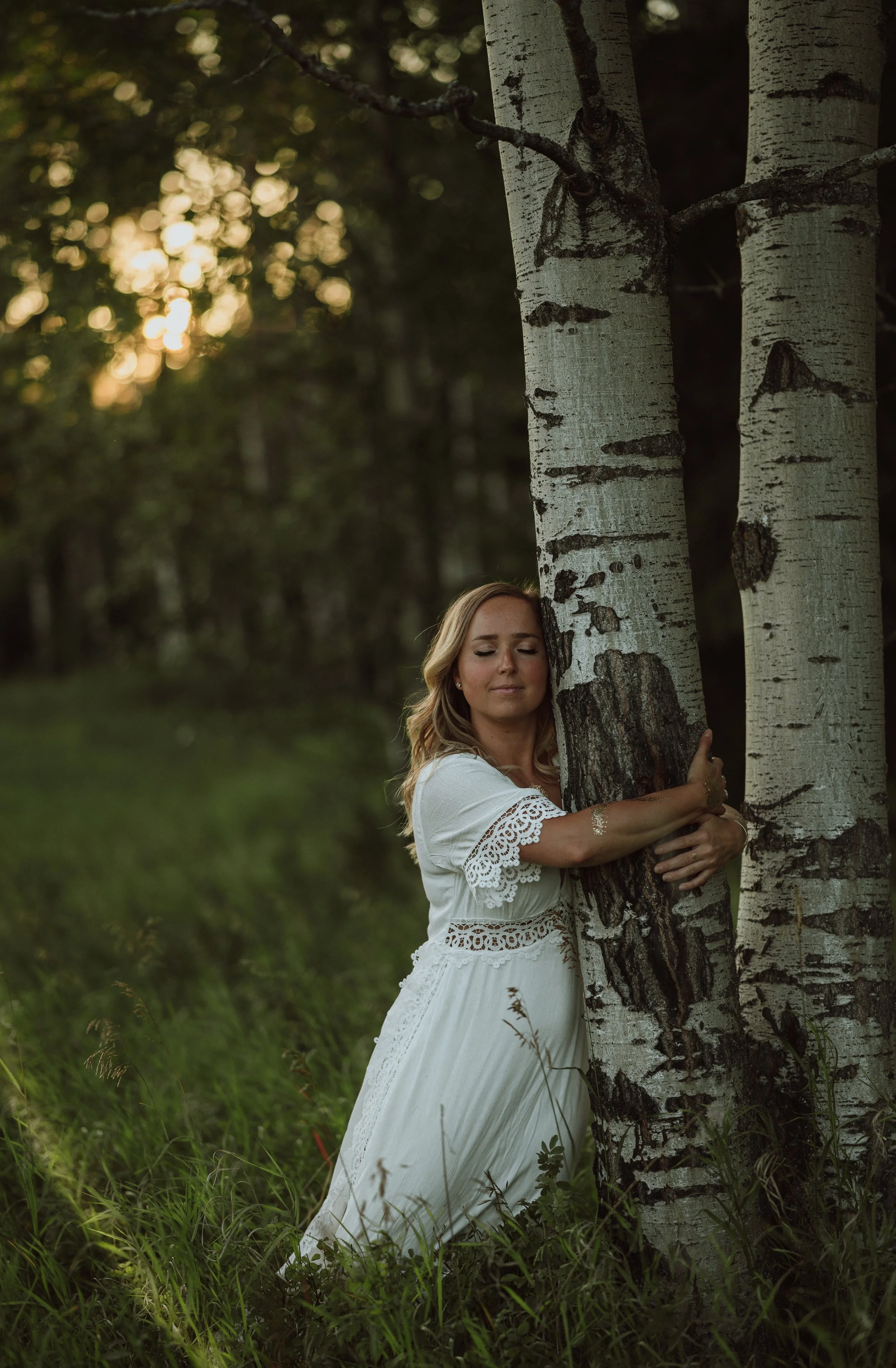 A woman in a white dress hugging a tree in a forested area during sunset with greenery and trees in the background.