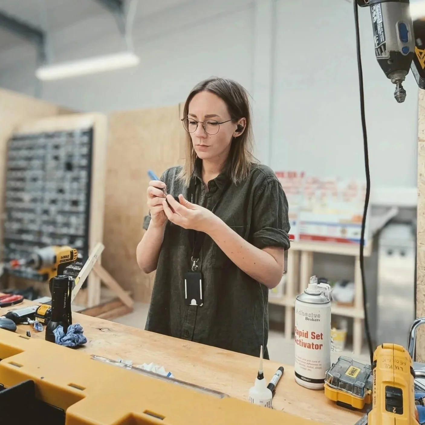 A woman with glasses and long hair, wearing a black shirt, standing in a workshop, holding a small object, surrounded by tools, a glue spray, and electrical equipment.