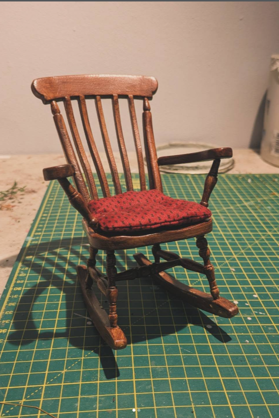 Miniature wooden rocking chair with a red cushioned seat, placed on a green cutting mat on a work surface.