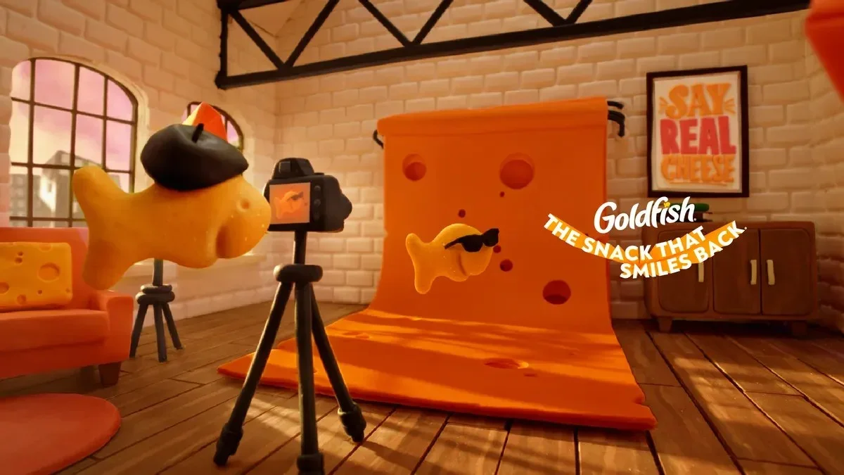 A plush cheese character with sunglasses being filmed in a cozy room with a brick wall, a window showing pink sky, an orange sofa, and a framed poster that says 'Say Real Cheese'.