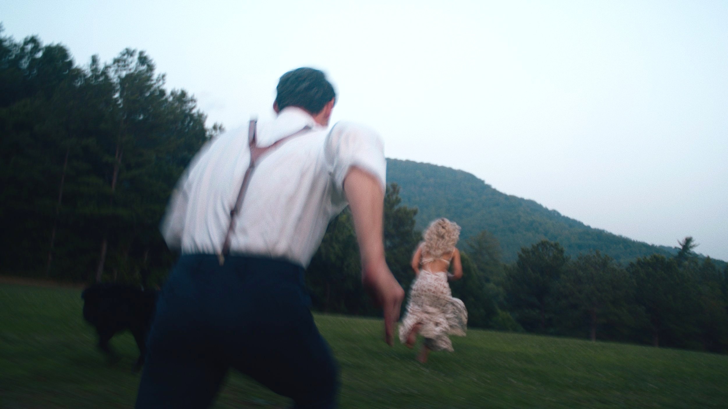 A man chasing a woman running across a grassy field with trees and mountains in the background.
