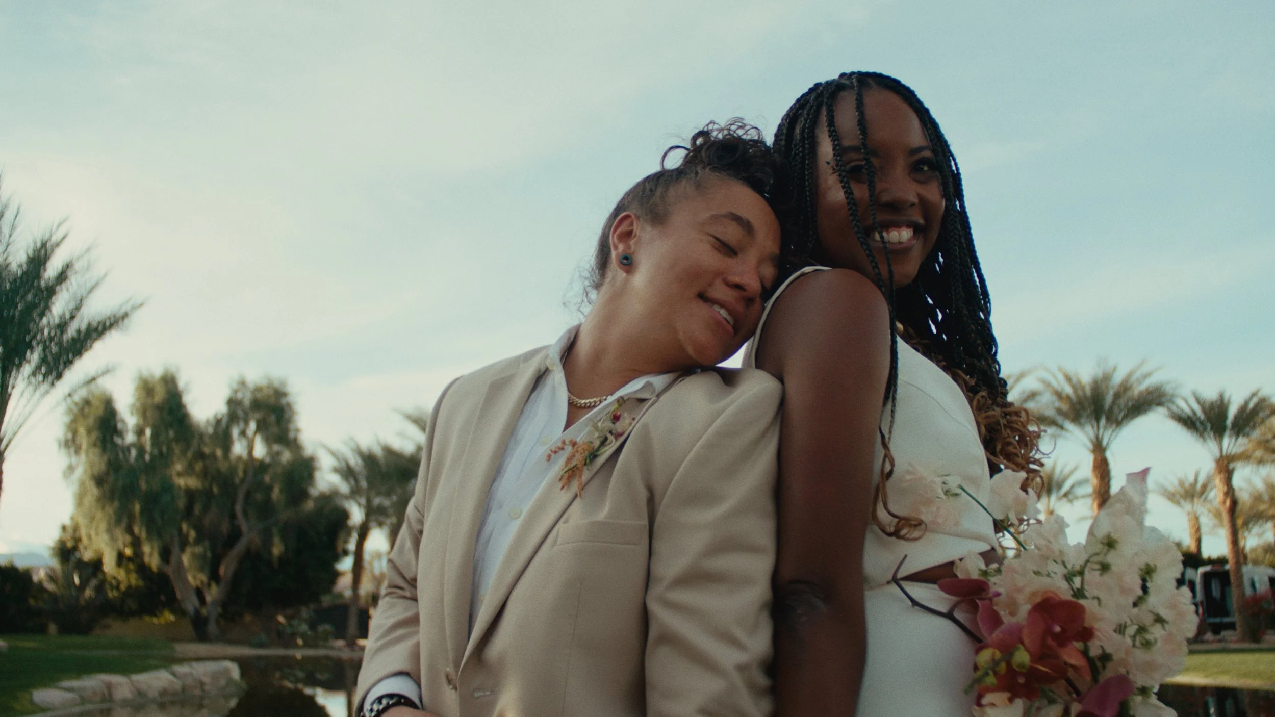 Two women smiling, one with short dark hair in a beige suit and the other with long braided hair holding a bouquet of flowers, outdoors with palm trees in the background.