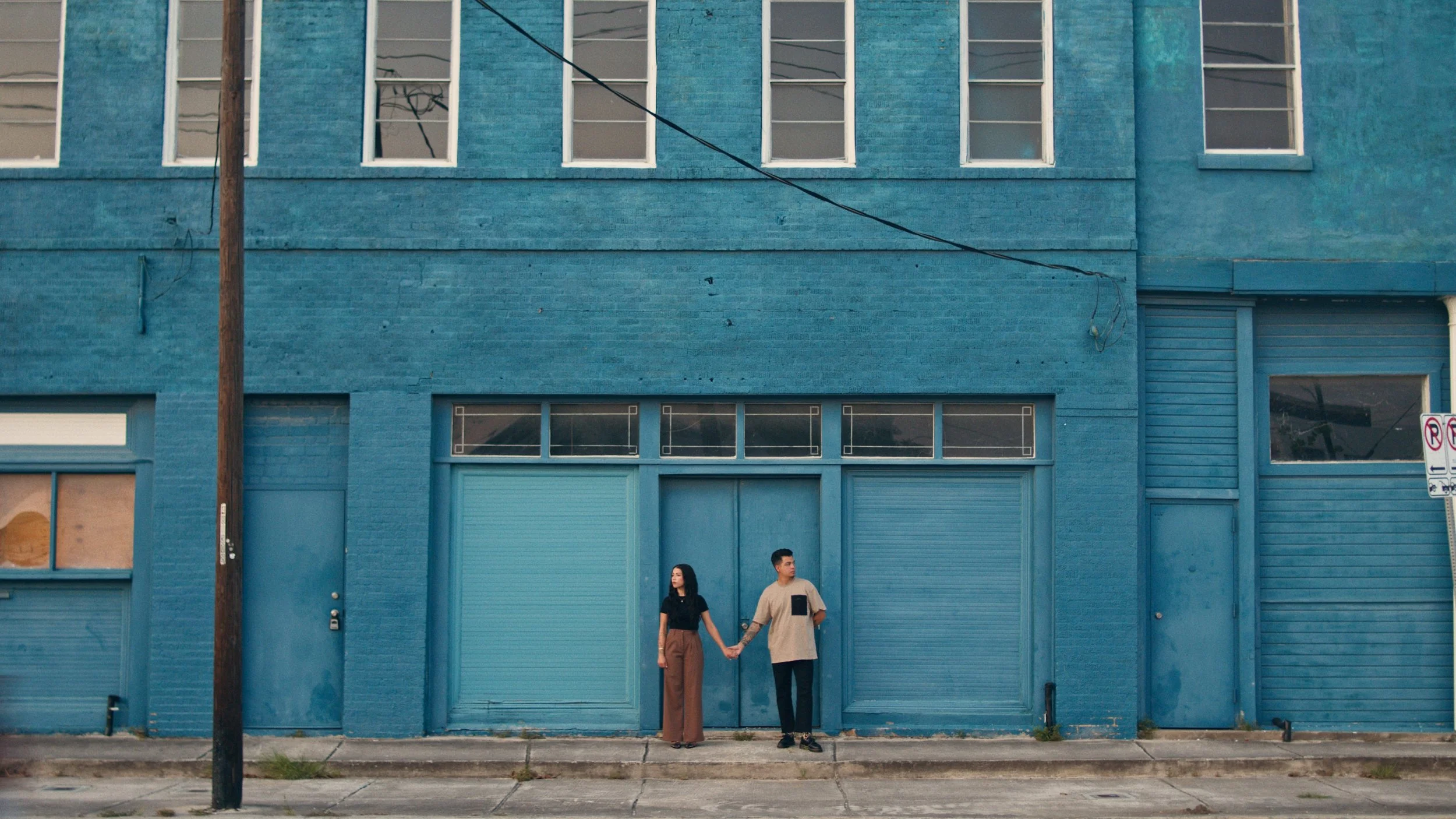 A man and woman holding hands stand in front of a large blue building with closed garage doors, windows, and a street with a pole and parking signs.